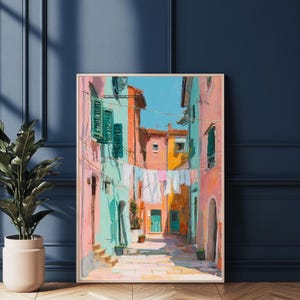 May include: A framed painting depicts a colorful Italian street scene. Buildings in shades of pink, turquoise, and orange line the narrow alleyway. White laundry hangs between the buildings, and a bright blue sky is visible above.