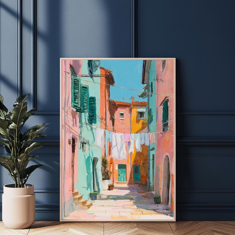 May include: A framed painting depicts a colorful Italian street scene. Buildings in shades of pink, turquoise, and orange line the narrow alleyway. White laundry hangs between the buildings, and a bright blue sky is visible above.