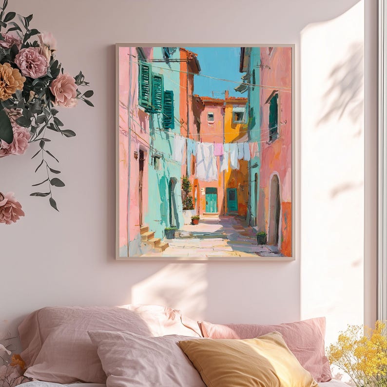 May include: A framed art print depicts a colorful Italian street scene. Buildings in shades of pink, turquoise, and orange line the narrow alleyway. Laundry hangs overhead, and a bright blue sky is visible. The artwork is a vibrant, impressionistic style.
