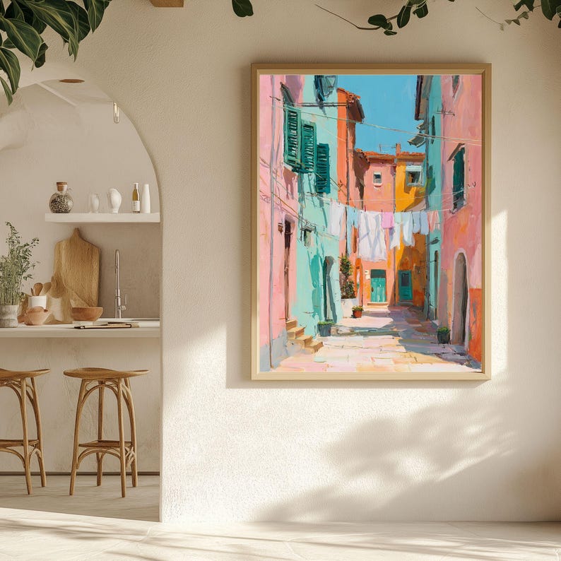 May include: A framed art print depicts a colorful street scene. Buildings in shades of pink, turquoise, and orange line the narrow street. Laundry hangs between the buildings under a bright blue sky. The artwork is in a light wood frame.