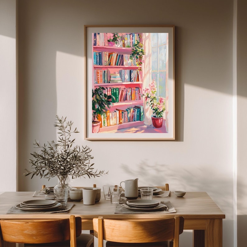 K&ouml;nnte beinhalten: Ein gerahmtes Gem&auml;lde zeigt ein rosa B&uuml;cherregal voller bunter B&uuml;cher, Pflanzen und Sonnenlicht, das durch ein Fenster scheint. Das Kunstwerk wird &uuml;ber einem Holzesstisch mit Tellern, Tassen und einer Vase mit Gr&uuml;nzeug ausgestellt.
