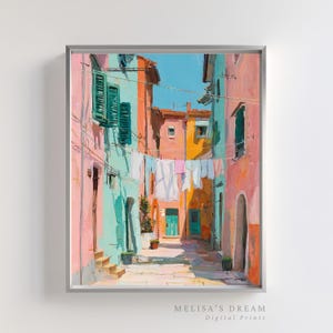 May include: A framed digital print of a colorful European street scene. The artwork depicts buildings in shades of pink, turquoise, and orange, with laundry hanging between them. The sky is a bright blue. The print is labeled "MELISA'S DREAM Digital Prints."