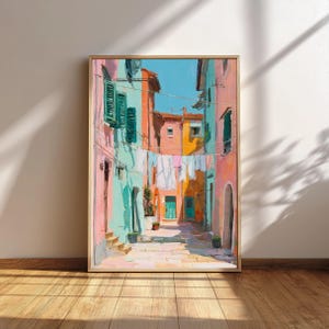 May include: A framed art print depicts a colorful Italian street scene. Buildings in shades of pink, turquoise, and orange line the cobblestone street. Laundry hangs between the buildings under a bright blue sky. The artwork is in a gold frame.