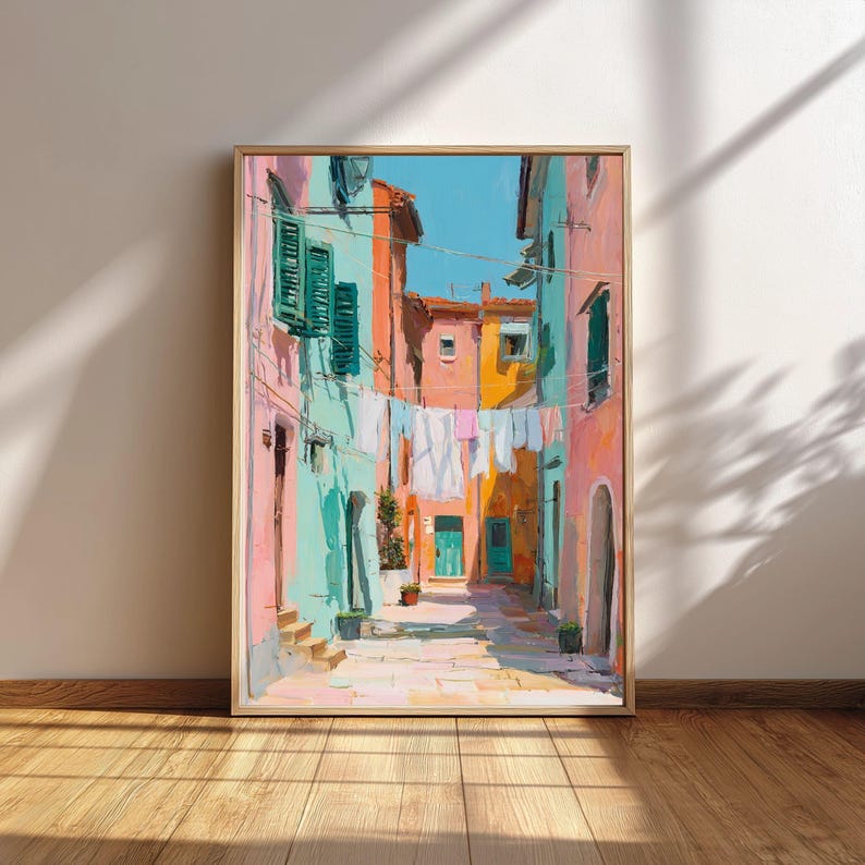 May include: A framed art print depicts a colorful Italian street scene. Buildings in shades of pink, turquoise, and orange line the cobblestone street. Laundry hangs between the buildings under a bright blue sky. The artwork is in a gold frame.