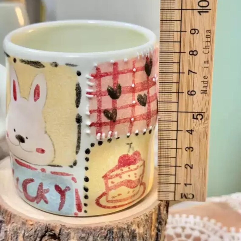 May include: A small, hand-painted ceramic mug with a whimsical design. The mug features a bunny, a cake, and the word "Lucky". The design includes red, yellow, and white colors. The mug is approximately 3 inches tall.
