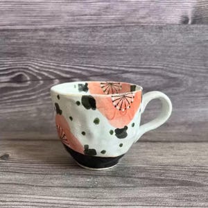 May include: A handmade ceramic mug with a white base, featuring a black rim and handle. The mug is decorated with coral-colored floral patterns and black and green abstract shapes. The mug is designed for hot beverages.