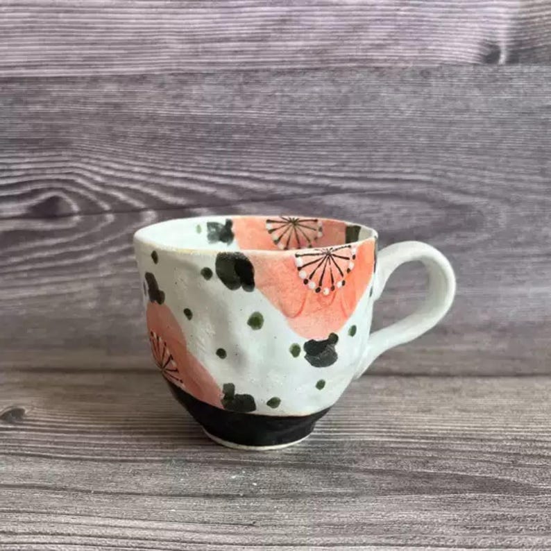 May include: A handmade ceramic mug with a white base, featuring a black rim and handle. The mug is decorated with coral-colored floral patterns and black and green abstract shapes. The mug is designed for hot beverages.
