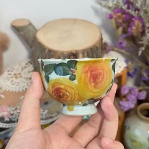 May include: A small, hand-painted ceramic bowl with yellow and red rose design. The bowl has a light blue base and a small pedestal. Green leaves and blue accents surround the roses. The bowl is held in a hand.