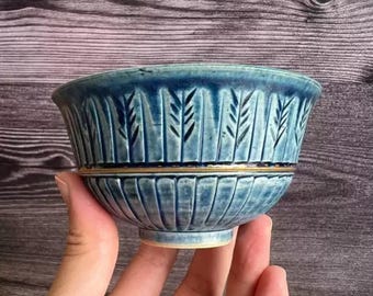 May include: A blue ceramic bowl with a textured surface and a gold band. The bowl features vertical grooves and leaf-like patterns. The bowl is held in a hand.