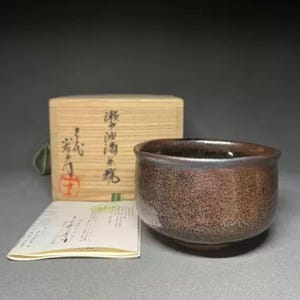 May include: A brown ceramic tea bowl with a textured surface. The bowl is next to a wooden box with Japanese characters and a small paper. The tea bowl has a dark interior and a rounded shape.