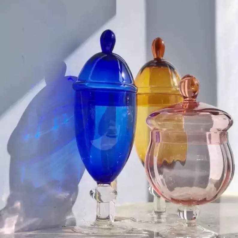May include: Three decorative glass jars with lids in blue, yellow, and pink. Each jar has a rounded body and a stem base. The lids have a small knob on top. The jars are transparent, allowing light to pass through.