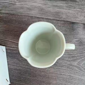 Puede incluir: Una taza de cer&aacute;mica verde claro con un borde &uacute;nico en forma de flor y un asa peque&ntilde;a. La taza tiene un acabado liso y brillante y est&aacute; dise&ntilde;ada para bebidas. La taza est&aacute; sobre un fondo de madera.