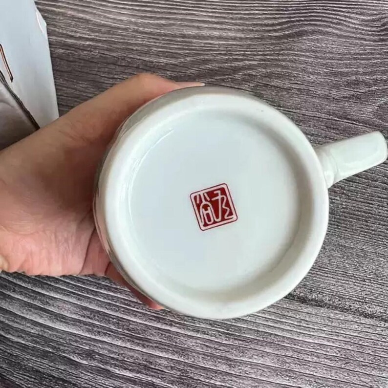 Puede incluir: Taza de cer&aacute;mica blanca con asa, con un sello cuadrado rojo en la base. La taza tiene un acabado liso y brillante. El asa es curvada y se extiende desde el lateral de la taza. La taza es probablemente para bebidas.