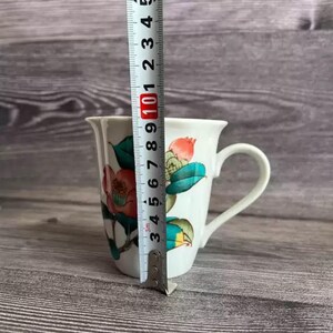 Puede incluir: Taza de cer&aacute;mica blanca con un dise&ntilde;o floral con flores y hojas rojas y verdes. La taza mide aproximadamente 10 cm de alto, medidos con una regla. La taza tiene un asa curva y un borde acampanado.