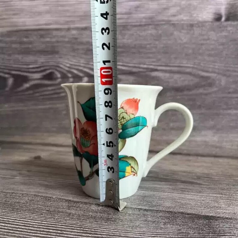 Puede incluir: Taza de cer&aacute;mica blanca con un dise&ntilde;o floral con flores y hojas rojas y verdes. La taza mide aproximadamente 10 cm de alto, medidos con una regla. La taza tiene un asa curva y un borde acampanado.