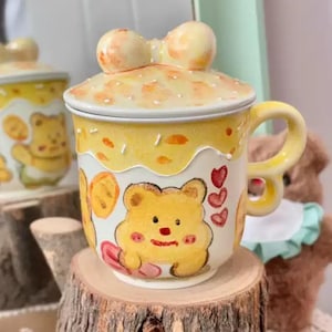 Puede incluir: Una taza de cerámica amarilla con tapa, con un diseño de oso de dibujos animados. La taza tiene un asa en forma de lazo y una tapa con un lazo en la parte superior. El diseño incluye corazones y un borde blanco. La taza está sobre una superficie de madera.