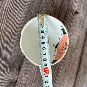 May include: A small, white ceramic bowl with a floral design in pink and black. The bowl is being measured with a ruler, showing a diameter of approximately 4 inches. The bowl sits on a wooden surface.