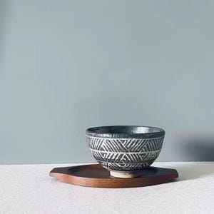 May include: A black and white patterned ceramic bowl sits on a wooden tray. The bowl has a geometric design and a dark interior. The tray is a dark brown color and has a unique shape. The background is a solid light blue color.