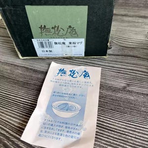 May include: A black box with Japanese text and a green label. A small white paper with Japanese text and a blue illustration of a bowl with two objects inside is in front of the box. The box and paper are on a wooden surface.