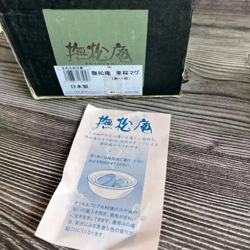 May include: A black box with Japanese text and a green label. A small white paper with Japanese text and a blue illustration of a bowl with two objects inside is in front of the box. The box and paper are on a wooden surface.