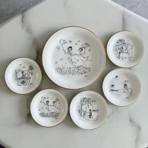 Puede incluir: Un juego de platos y platillos de cerámica blanca con bordes dorados. Cada pieza presenta una ilustración en blanco y negro de personas en varias escenas, incluyendo parejas bailando y músicos tocando. El juego incluye un plato grande y varios platillos.
