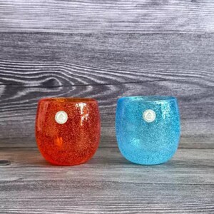 Puede incluir: Dos candelabros votivos de cristal brillantes, uno naranja y otro azul, uno al lado del otro. Cada uno tiene una apariencia texturizada y brillante y un pequeño emblema redondo plateado. Los candelabros están sobre una superficie de madera.