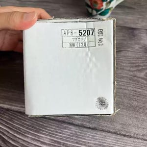 Puede incluir: Una caja de cart&oacute;n blanca con texto japon&eacute;s y una pegatina. La caja tiene el texto "AP6-5207" y "マグカップ" impreso. La caja est&aacute; sujeta por una mano.