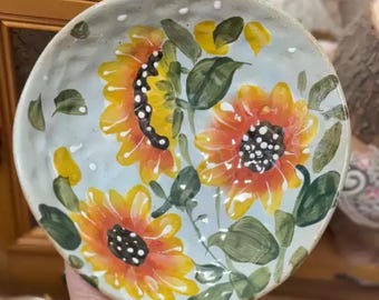 May include: Hand-painted ceramic bowl featuring vibrant sunflowers in shades of yellow and orange with black centers. The bowl has a light blue background with small white dots and green leaves. A decorative piece for the kitchen or dining room.