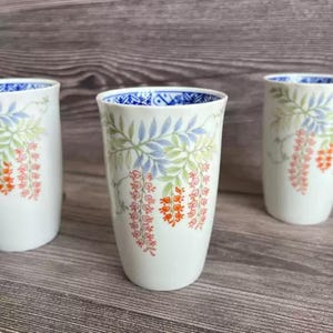 Puede incluir: Tres vasos de cerámica blanca con un diseño floral. Los vasos presentan un borde azul y un patrón de hojas verdes, ramas azules y flores naranjas. Los vasos están colocados sobre un fondo de madera.