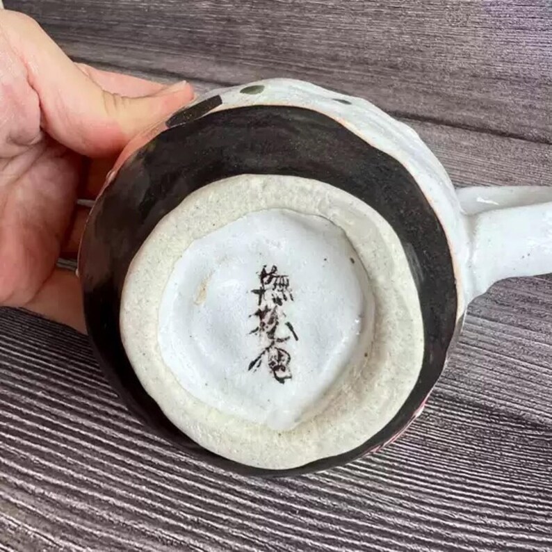 May include: A handmade ceramic mug with a white handle and a black and white design. The bottom of the mug is white with black Japanese calligraphy. The mug is held by a hand.