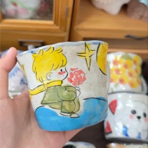 Könnte beinhalten: Handbemalte Keramiktasse mit einer skurrilen Illustration eines Charakters, der eine Blume hält. Die Tasse hat einen blauen Boden, einen gelben Stern und Mond sowie eine Figur mit gelben Haaren, grünem Mantel und gelbem Schal.