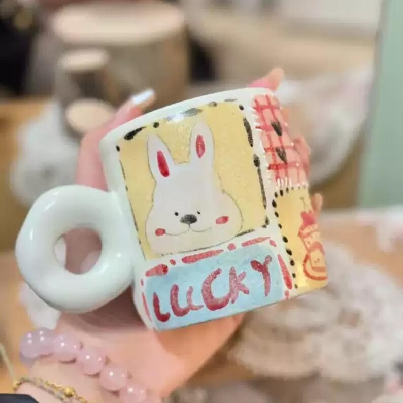 May include: A white ceramic mug with a rounded handle. The mug features a hand-painted design of a white bunny with red ears and cheeks, and the word "LUCKY" in blue. The mug has a yellow background with black border.
