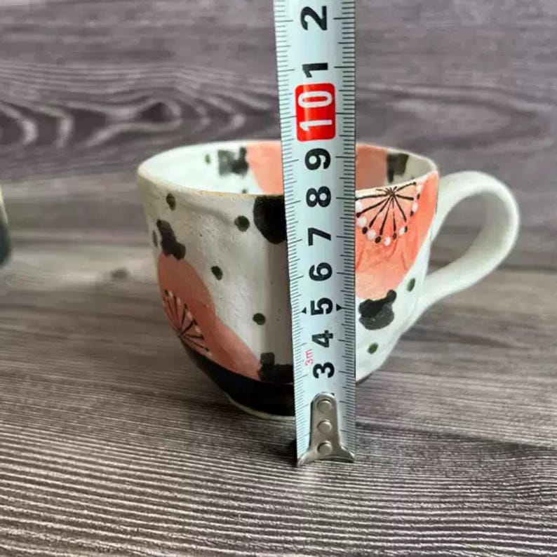 May include: A white ceramic mug with a handle, decorated with black polka dots and coral-colored floral designs. The mug is approximately 4 inches tall, as measured by a tape measure.