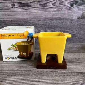 Può includere: Una fonduta gialla con una base in legno marrone scuro. La pentola ha una forma quadrata con manici sui lati. La scatola accanto dice "fondue set Tapas" e presenta il marchio "BOSKA".