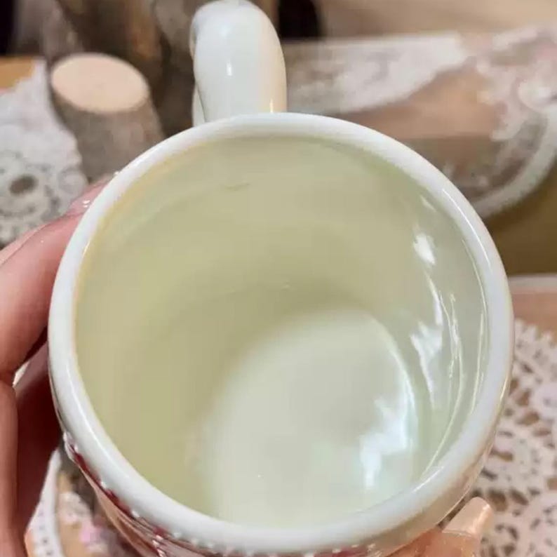 May include: Cream-colored ceramic mug with a curved handle. The interior of the mug is a pale cream color. The rim of the mug has a decorative pattern in red. The mug is held by a person.