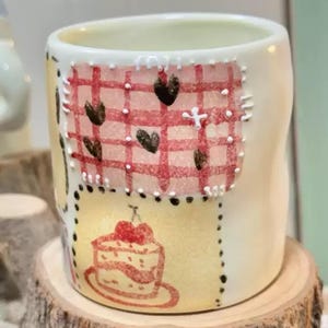 May include: A white ceramic mug with hand-painted designs. The mug features a red and white checkered pattern with black hearts, a yellow patch with a cake illustration, and black dotted borders. The mug is placed on a wooden surface.