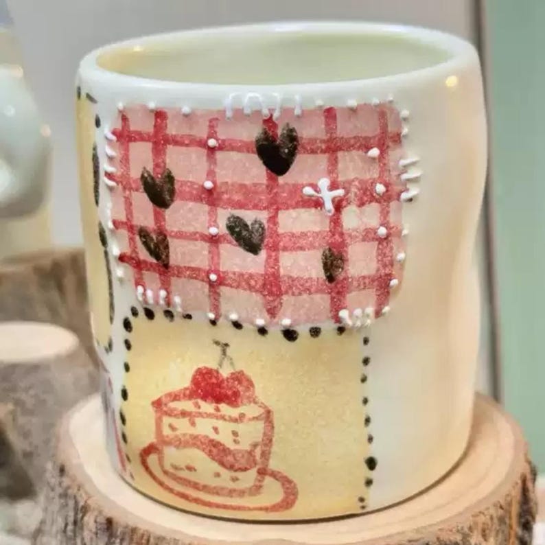 May include: A white ceramic mug with hand-painted designs. The mug features a red and white checkered pattern with black hearts, a yellow patch with a cake illustration, and black dotted borders. The mug is placed on a wooden surface.