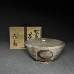 May include: A handmade ceramic bowl with a textured, off-white and brown glaze. A dark brown circle design is painted on the side. A wooden box with Japanese characters is in the background.