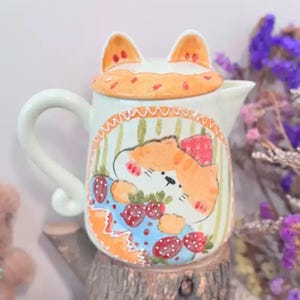 May include: A decorative ceramic teapot featuring a cat design. The teapot is white with a lid shaped like cat ears and a curved handle. The body of the teapot has a hand-painted illustration of a cat with strawberries. The cat is orange and white, and the strawberries are red.