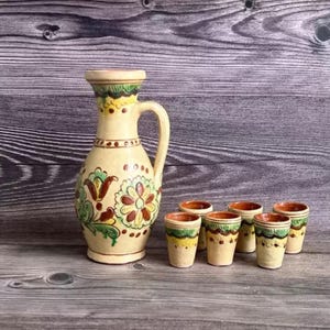Puede incluir: Una jarra de cerámica color crema con asa y diseños florales en verde, marrón y amarillo. Seis pequeñas tazas a juego con patrones similares están dispuestas al lado. El conjunto está sobre una superficie de madera.