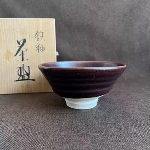 May include: A dark brown ceramic bowl with a white base. The bowl has a textured, ridged exterior and a smooth, glossy interior. A wooden box with Japanese characters is partially visible in the background.