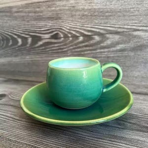 Puede incluir: Un pequeño juego de taza y platillo de cerámica hecho a mano. La taza y el platillo son de un verde vibrante con una superficie texturizada. El interior de la taza tiene un esmalte azul claro. El juego está diseñado para servir té o café.
