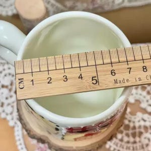 May include: A white ceramic mug with a handle, measuring approximately 2 inches in diameter, is shown with a wooden ruler. The mug has a painted design around the base. The ruler is marked in centimeters.