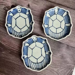 May include: Three ceramic plates with a turtle design. The plates are white with a blue turtle illustration. The turtle's shell is a geometric pattern, and the plates have a scalloped edge.