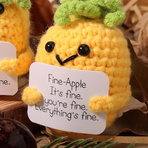 Puede incluir: Una piña amarilla tejida con ojos negros y una parte superior verde. La piña sostiene un letrero blanco que dice "Fine-Apple It's fine. you're fine. Everything's fine."
