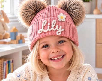 Puede incluir: Un gorro de punto acanalado rosa con dos grandes pompones de piel sintética marrón en la parte superior, un detalle de flor bordada en blanco y el nombre Lily cosido en letras cursivas blancas al frente.