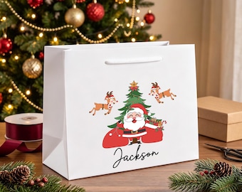 Op de afbeelding: Een witte cadeauzak met een afbeelding van de Kerstman voor een kerstboom met twee rendieren, en de naam Jackson in zwarte schrijfletters onder de illustratie. De tas heeft witte handvatten.