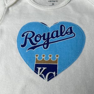 Pu&ograve; includere: Body da bambino bianco con toppa blu a forma di cuore. La toppa presenta la scritta "Royals" in corsivo, una corona e le lettere "KC" in un design con logo della squadra. Il body &egrave; taglia 6.