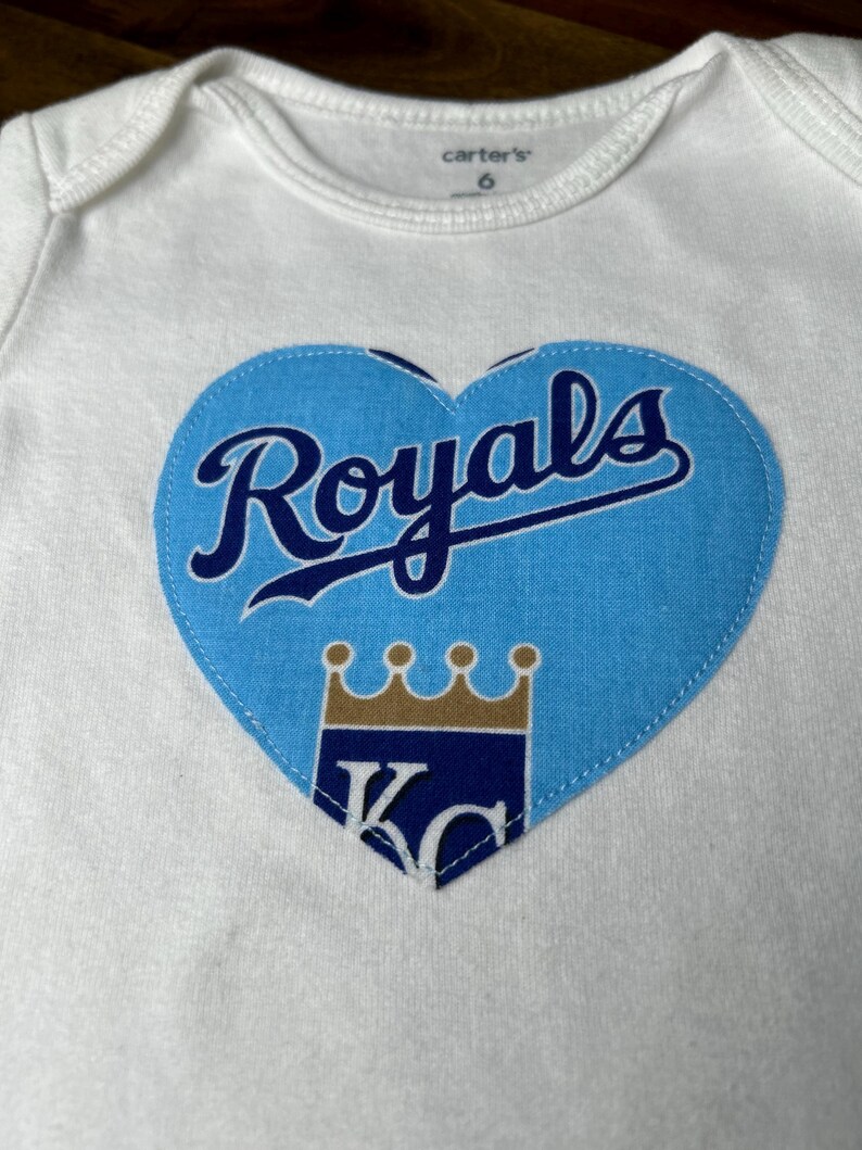 Pu&ograve; includere: Body da bambino bianco con toppa blu a forma di cuore. La toppa presenta la scritta "Royals" in corsivo, una corona e le lettere "KC" in un design con logo della squadra. Il body &egrave; taglia 6.
