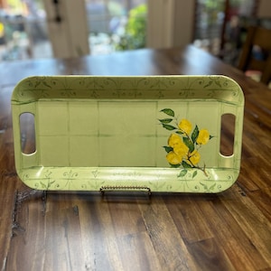 May include: Rectangular serving tray with a light green background and a lemon branch design. The tray has cut-out handles and decorative scrollwork. The lemons are yellow with green leaves. The tray is on a wooden surface.